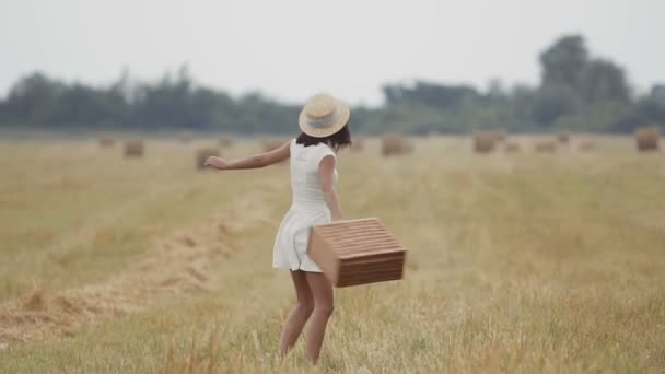 Ein junges Mädchen mit Strohhut und weißem Kleid läuft fröhlich über das Feld. hält das Mädchen einen Strohkorb in den Armen. Sonnenuntergang — Stockvideo