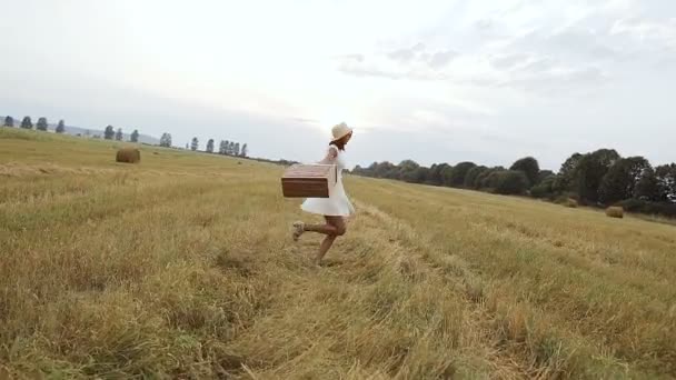 フィールド。日没。若い女の子は、麦藁帽子とスーツケースを持つフィールドに行く. — ストック動画