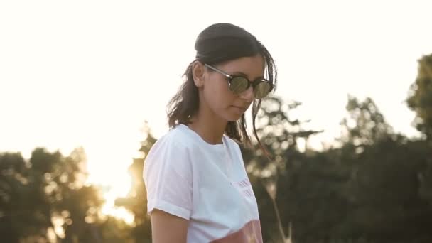 Молода жінка в сонцезахисних окулярах на заході сонця стоїть на пшеничному полі — стокове відео