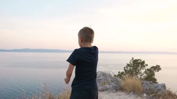 El niño está parado en una montaña de acantilados y arroja piedras al mar desde la altitud. Puesta de sol — Vídeos de Stock