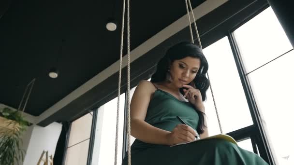 Jovem bela mulher sentada em um balanço e fazendo gravações em seu caderno — Vídeo de Stock
