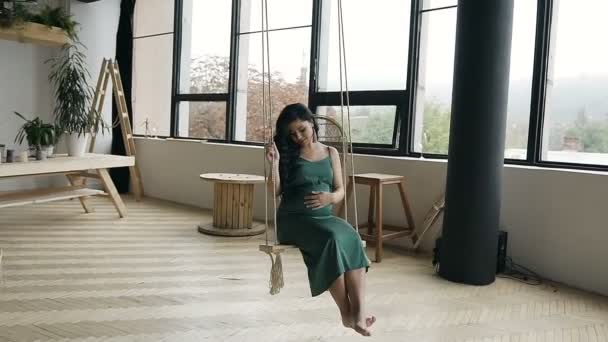 Giovane ragazza incinta oscillante su un'altalena di legno. Una giovane ragazza in un lungo vestito verde su un'altalena in studio . — Video Stock