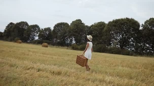 Mladá dívka, která nosí bílé šaty s slaměný klobouk a kufr. Sláma a slaměné balíky. Západ slunce — Stock video