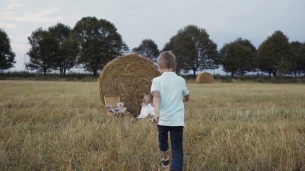 Een kleine jongen loopt naar een meisje in het veld onder de baal stro zit. Gelukkig meisje in witte jurk lachende jongen loopt naar haar. Zonsondergang — Stockvideo