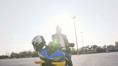 Bir motorcu bir deri ceket ve güneş gözlüklü, gün batımında, motosiklet yanında duruyor ve telefonla konuşuyor