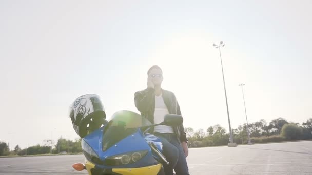 Motociclista con chaqueta de cuero y gafas de sol, se para junto a su moto y habla por teléfono — Vídeo de stock