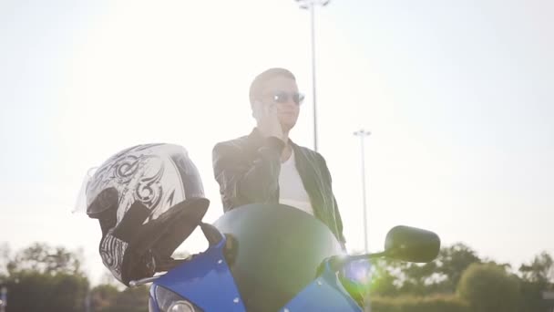 Een jonge man in een leren jas en zonnebril zittend op zijn motorfiets en plezier praten aan de telefoon — Stockvideo