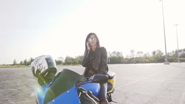 En ung biker flicka med mörkt hår i en skinnjacka och solglasögon sitter på sin motorcykel och har kul att prata i telefon — Stockvideo
