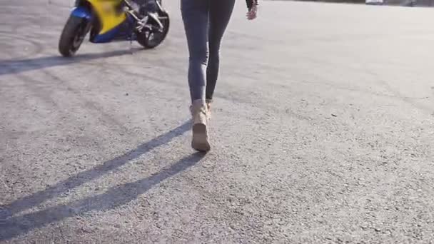 Mladá dívka má batoh na ramena, ona je oblečená v džínách a koženou bundu a ona se blíží motocyklu a na něm sedí — Stock video