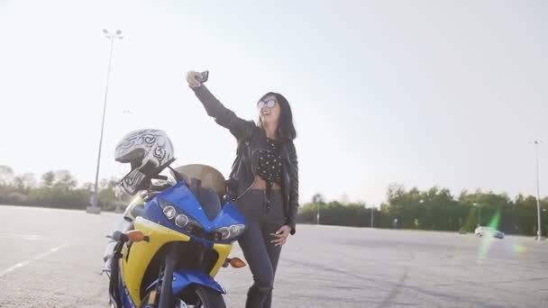 Libertad y estilo. La chica motociclista se para junto a su motocicleta amarilla y azul y felizmente hace un sephi en su teléfono. La chica sostiene el teléfono en su mano — Vídeo de stock