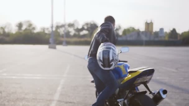 Un joven motociclista vestido con una chaqueta de cuero negro y vaqueros azules con un casco en las manos va a su motocicleta — Vídeos de Stock