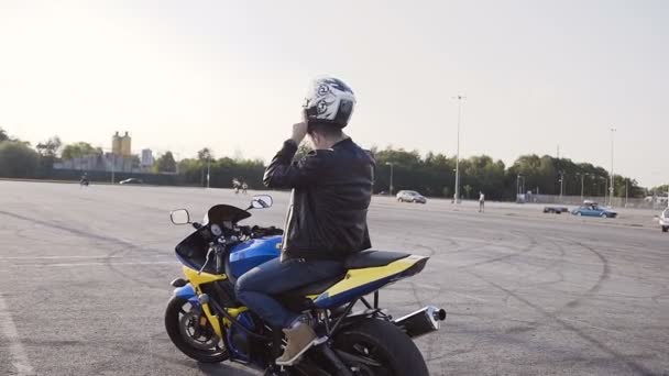Motocyklista na západ slunce sedne si na motorce, nosí helmu a jde do města — Stock video
