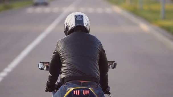 Un joven motorista con una chaqueta de cuero y un casco a caballo en la motocicleta a lo largo de la carretera alrededor de la ciudad. Primer plano — Vídeo de stock
