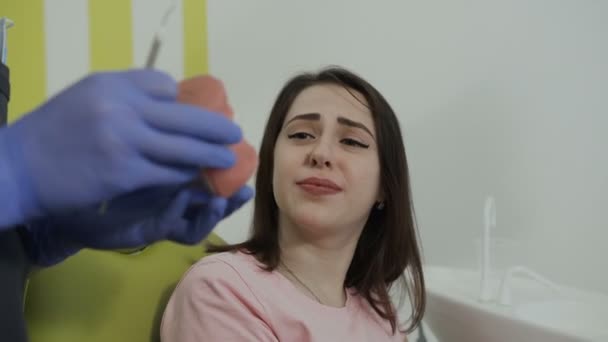 Hermosa paciente joven está sonriendo mientras se sienta en la silla en el dentista. Dentista guapo mostrando mandíbula artificial en el consultorio dental — Vídeos de Stock