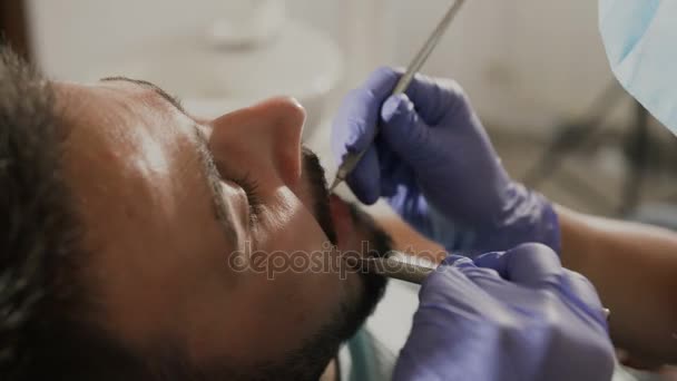 Female dentist examining patient teeth with a mouth mirror and dental excavator. Close-up view on the mans face — Stock Video