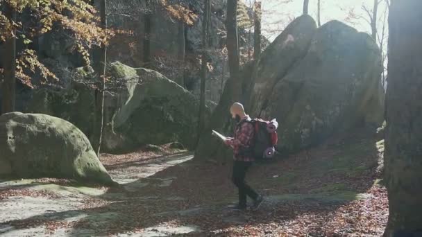 Hipster vestido con jeans negros y una camisa en una jaula camina por el bosque donde se encuentran las rocas y sostiene una mochila sobre sus hombros y una tarjeta turística en sus manos. Otoño — Vídeos de Stock