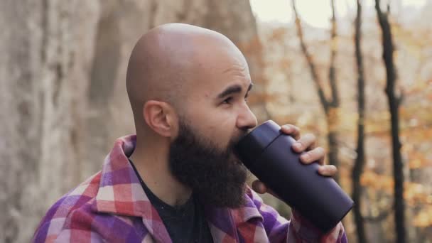 Hermoso hombre barbudo se sentó a descansar en el bosque cerca de la roca, sosteniendo un termo en sus manos y bebiendo té de él — Vídeos de Stock