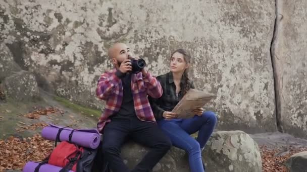 Ein junger Reisefotograf mit Kamera, Touristenkarte und Rucksack sitzt mit einem jungen Mädchen auf einem Felsen und blickt auf den Sonnenuntergang — Stockvideo