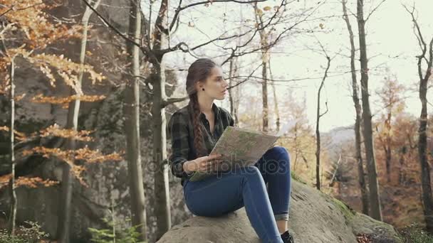 Belle jeune fille aux cheveux longs tressée dans une tresse, assise sur un rocher dans les bois et regardant la carte. Automne doré — Video