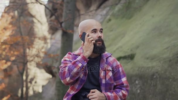 Primer plano. Joven sentado en una roca cerca de una roca alta y hablando en un teléfono móvil en una ciudad turística. Maravilloso otoño — Vídeos de Stock