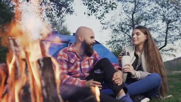 Feliz pareja joven sentada junto al fuego en un pueblo de tiendas de campaña. Los viajeros descansan con una taza de té caliente en el bosque cerca de una tienda azul — Vídeo de stock