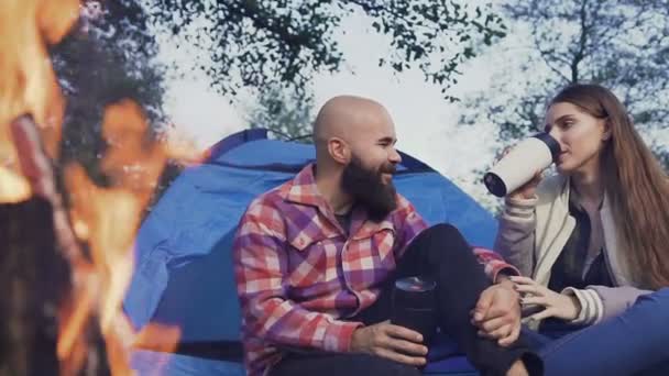 Feliz pareja joven sentada junto al fuego en un pueblo de tiendas de campaña. Los viajeros descansan con una taza de té caliente en el bosque cerca de una tienda azul — Vídeos de Stock