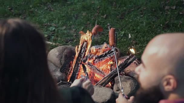 Griller des saucisses sur un feu de camp, des campeurs rôtissant des saucisses. Les touristes sont assis près de la flamme — Video