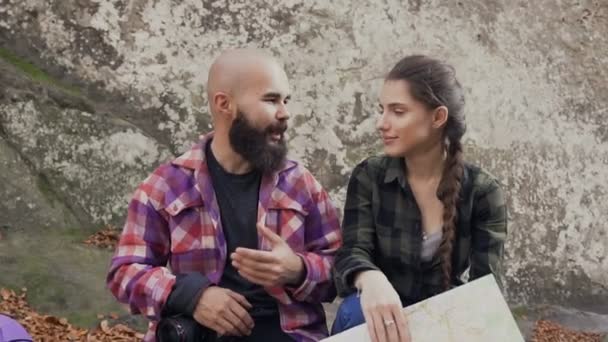 Un bel giovane uomo con la barba parla gioiosamente con una bella ragazza. I turisti si sono seduti a riposare sulla pietra e parlare della loro ulteriore escursione in montagna. — Video Stock
