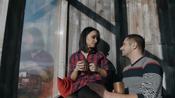 Gyönyörű fiatal pár ül körül a ablak otthon egy napsütéses téli nap, és italt forró csokoládé. Karácsony és nyaralás — Stock videók
