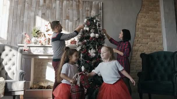 Los padres jóvenes decoran un árbol de Navidad, y sus hijas gemelas montan un caballo de juguete. Feliz Año Nuevo y Navidad — Vídeo de stock