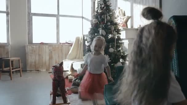 Duas meninas pequenas morena e loira correm em torno de uma poltrona macia perto de uma árvore de Natal na véspera de Natal — Vídeo de Stock