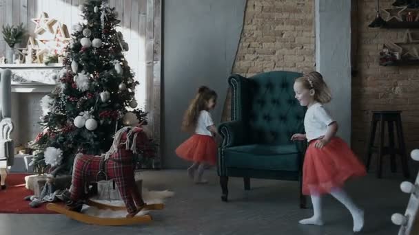 Two little girls - brunette and blonde run around a soft armchair near a Christmas tree on Christmas Eve — Stock Video