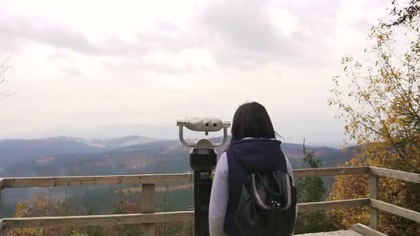 Chica viendo el horizonte. Una joven turista con mochila oscura se adapta a los prismáticos de pie en la plataforma de observación en las montañas. Binocular en la montaña, moneda operado binocular — Vídeos de Stock