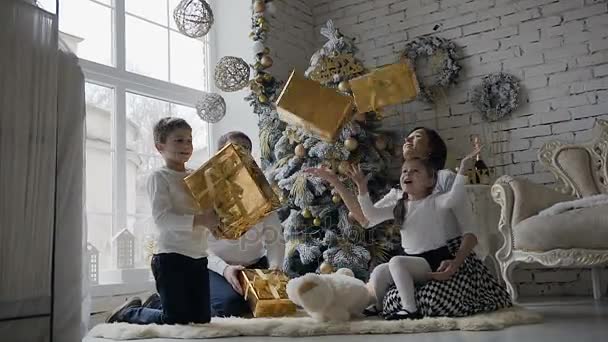 Νεαρή οικογένεια ντυμένος στα λευκά ρούχα, καθίστε στο πάτωμα κοντά στο χριστουγεννιάτικο δέντρο στο σαλόνι με ένα μεγάλο παράθυρο και χαίρονται με τα δώρα που βρίσκονται στην Χρυσή κουτιά — Αρχείο Βίντεο