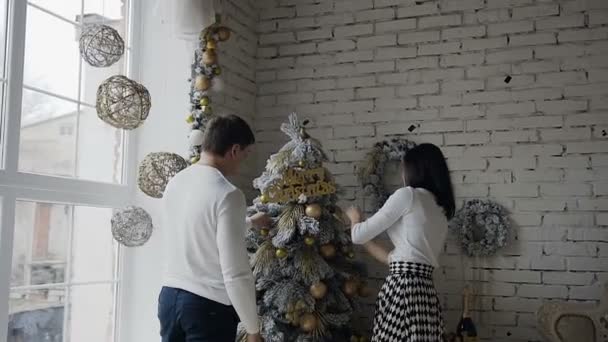 Ein Mann mit einer Frau wird mit einem Weihnachtsbaum geschmückt. eine glückliche Familie schmückt einen dekorativen Weihnachtsbaum neben einem großen Fenster im Wohnzimmer — Stockvideo