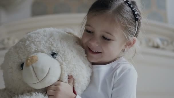 Gros plan. La petite fille s'assoit sur un tapis blanc au lit et étreint un jouet d'ours doux. Belle fille et son ours jouet doux blanc — Video