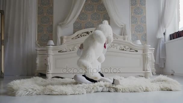 Очаровательная девушка сидит на полу у кровати и радостно играет с мягким плюшевым мишкой. Веселый малыш и ее плюшевый мишка мягкая, белая игрушка — стоковое видео