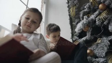 Yakın çekim çocuk Noel ağacının yakınında büyük bir pencere pencere pervazına oturmak ve ilginç kitap okumak