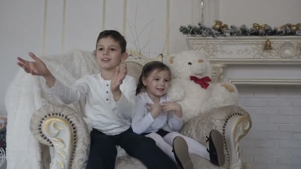 Des enfants heureux - un garçon et une fille sont assis sur une grande chaise et attrapent des cadeaux dans des boîtes dorées. Soirée de Noël — Video