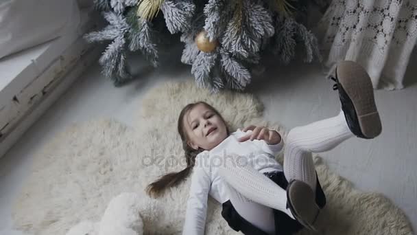 La encantadora chica se encuentra en una suave alfombra blanca y se juega con un gran oso de peluche. Vacaciones de invierno. Árbol de Navidad — Vídeos de Stock