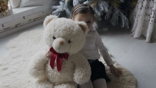 Hermoso bebé está jugando alegremente con un gran oso de peluche blanco en el suelo cerca del árbol de Navidad en la sala de estar. Hermosa chica en una alfombra blanca cerca del árbol de Navidad — Vídeos de Stock