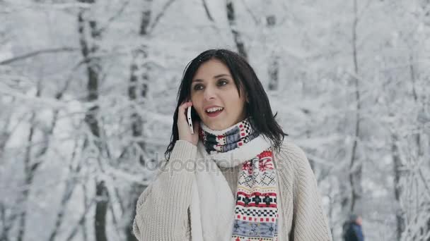 Una hermosa chica en el bosque de invierno habla por teléfono. Hermosa joven morena utiliza un teléfono inteligente, caminando en un parque cubierto de nieve en un suéter marrón de moda, bufanda caliente y mitones — Vídeos de Stock