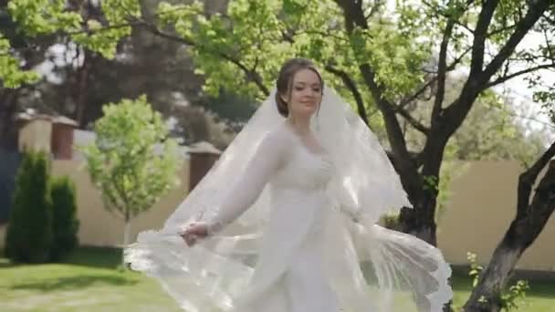 Gros plan. Belle mariée en robe de mariée fantastique avec une longue dentelle tournant autour et ayant des promenades dans le jardin de printemps près de sa maison — Video