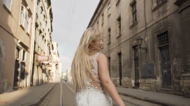 Belle jeune fille blonde en robe blanche aux cheveux longs traverse les rues de la ville le matin. Lever de soleil, charmante mariée — Video