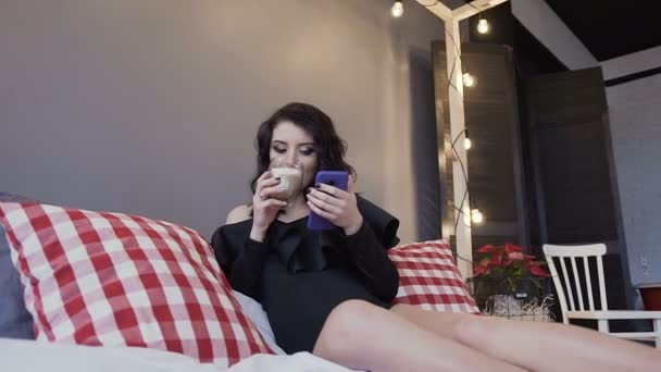 Счастливая беременная молодая женщина пьет теплый шоколад и пользуется сотовым телефоном дома — стоковое видео