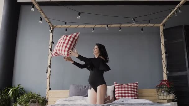 Zwangere brunette meisje, verheugt zich en gooit een kussen. Vrouw in bed in een zwarte zwembroek — Stockvideo