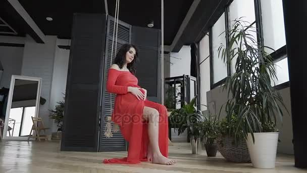 Dziewczyna w ciąży długo czerwona sukienka siedzi na huśtawce i delikatnie głaszcze jej brzuch i patrzy na niego. Poczęcie w ciąży. Szczęśliwy czas — Wideo stockowe