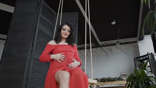 Беременная девушка в длинном красном платье сидит на качелях, нежно гладит живот и смотрит на него. Зачатие беременности. Happy time — стоковое видео
