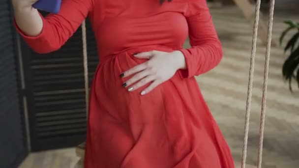 Κοντινό πλάνο. Μια έγκυος γυναίκα σε ένα κόκκινο μακρύ φόρεμα που μιλάτε στο τηλέφωνο μέσω του κοινωνικού δικτύου χρησιμοποιώντας το ασύρματο vaj αρχείων internet και δείχνοντας τα χέρια στην κοιλιά της — Αρχείο Βίντεο