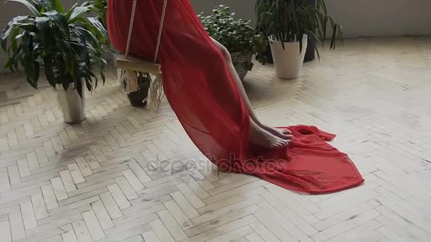 Young pregnant woman swinging on a wooden swing. A young girl in a long red dress on a swing in the studio — Stock Video